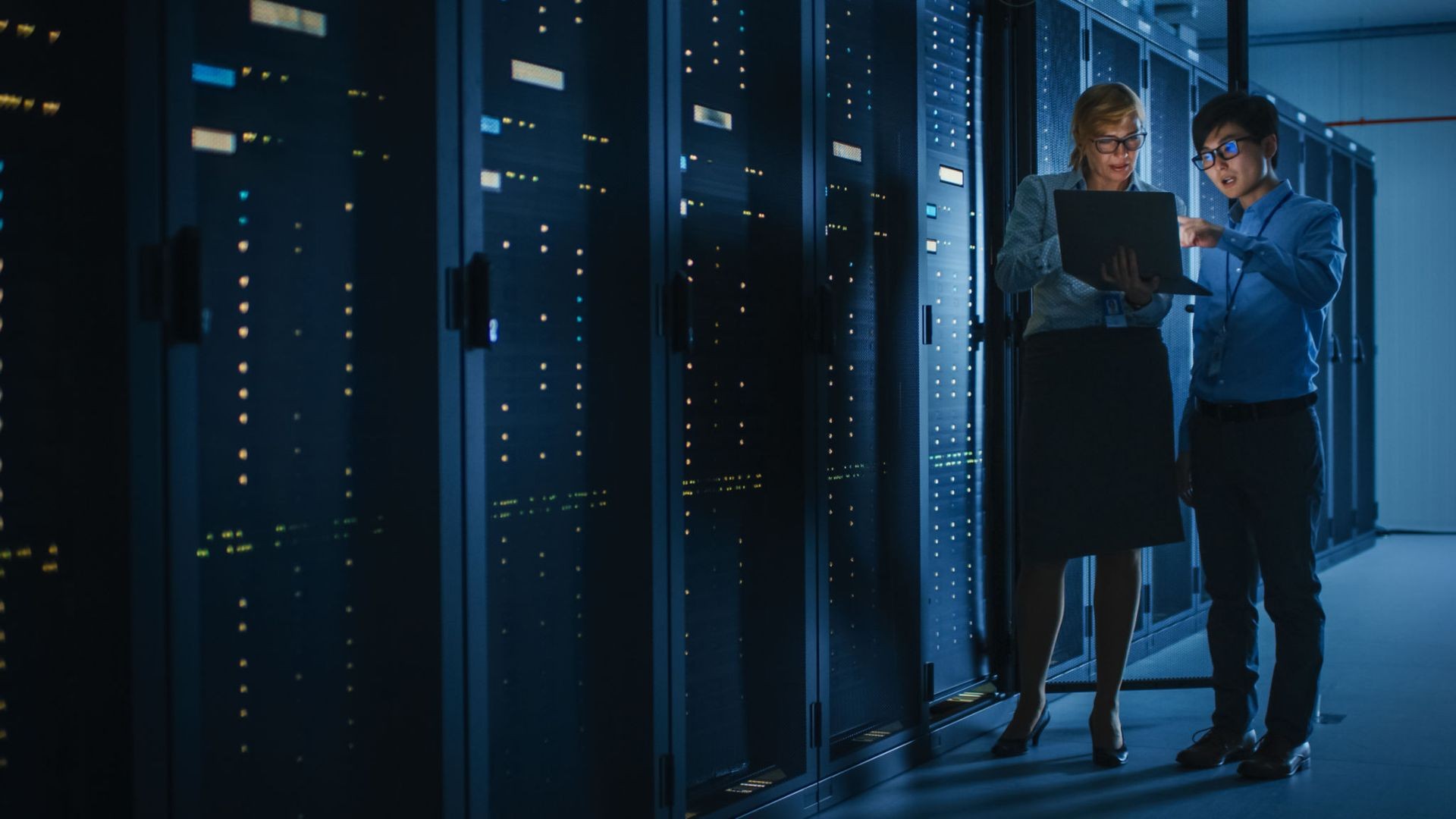 Two professionals discussing data on a laptop in a dimly lit server room.